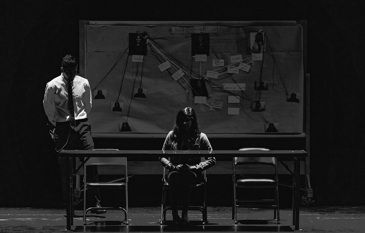 Een spannende, donkere foto met een vrouw aan tafel en een man in pak met een groot bewijsbord voor een moordonderzoek op de achtergrond. Op het bewijsbord zijn personen, aantekeningen en touwtjes die bewijsmaterialen verbinden te zien.